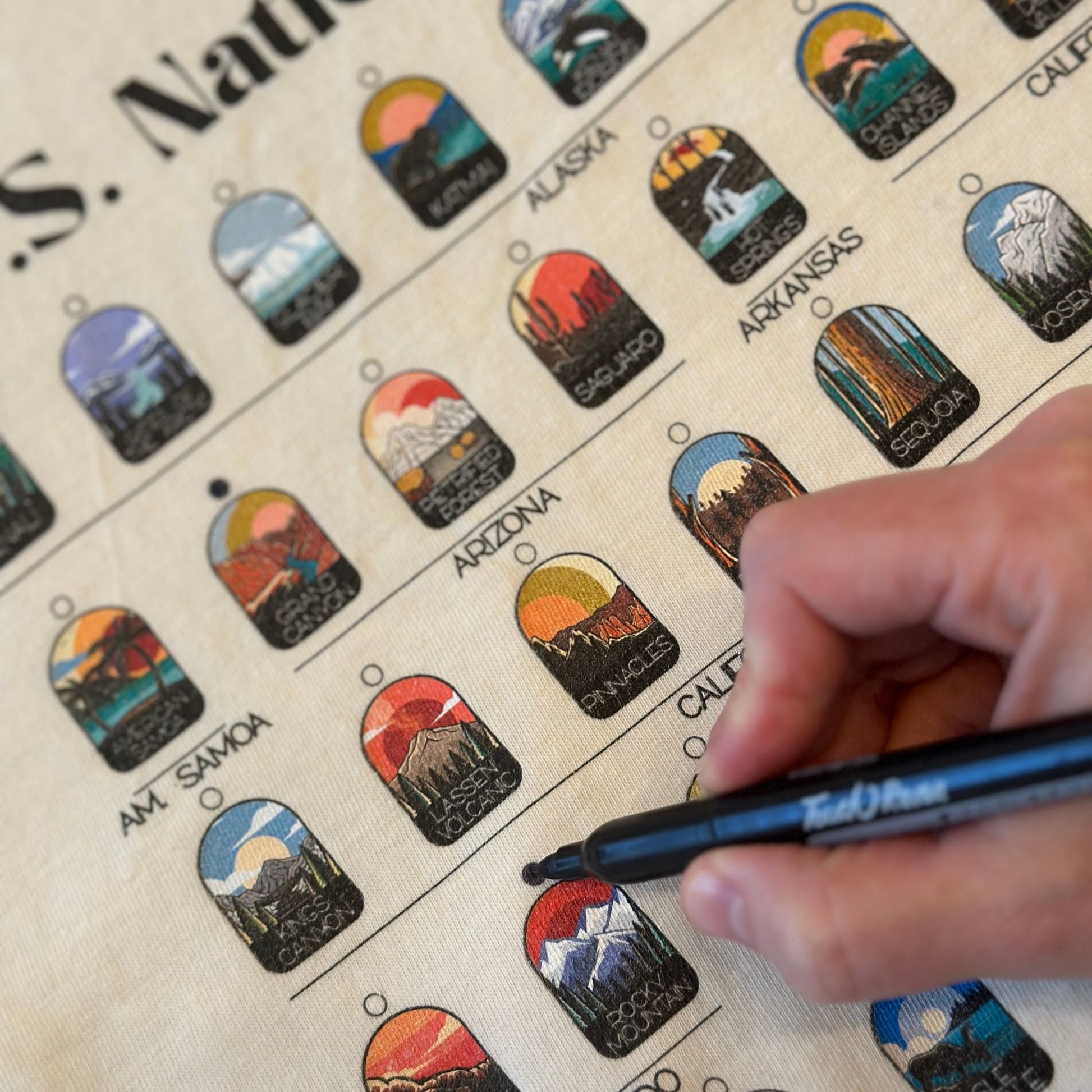 Cream t-shirt with all 63 National Parks. Someone is filling in the circles with a permanent marker to track your National Parks.