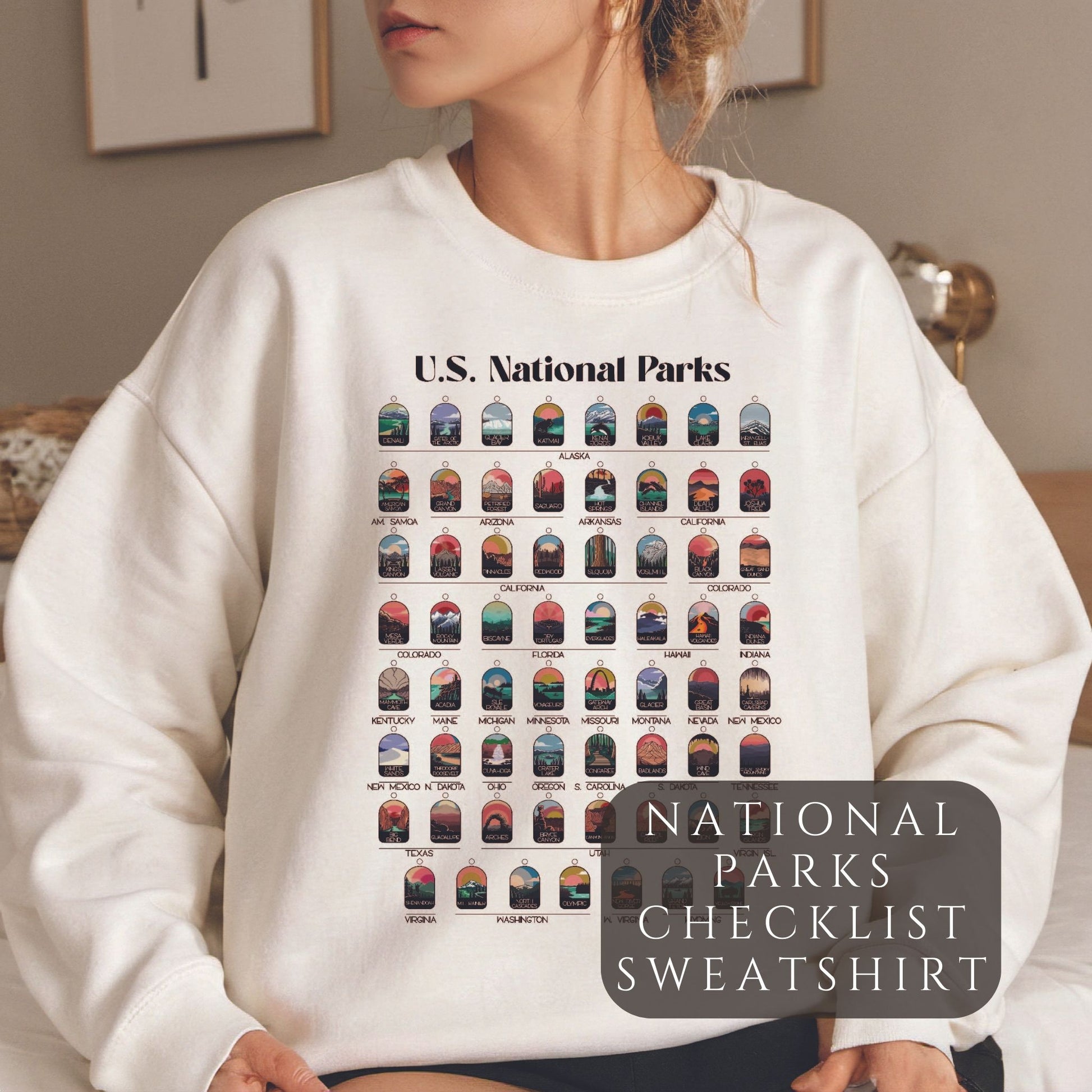 Woman wearing cream colored sweatshirt. Sweatshirt has all 63 National Parks on it with circles to fill in to track your National Parks.