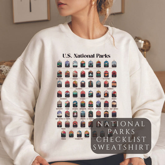 Woman wearing cream colored sweatshirt. Sweatshirt has all 63 National Parks on it with circles to fill in to track your National Parks.