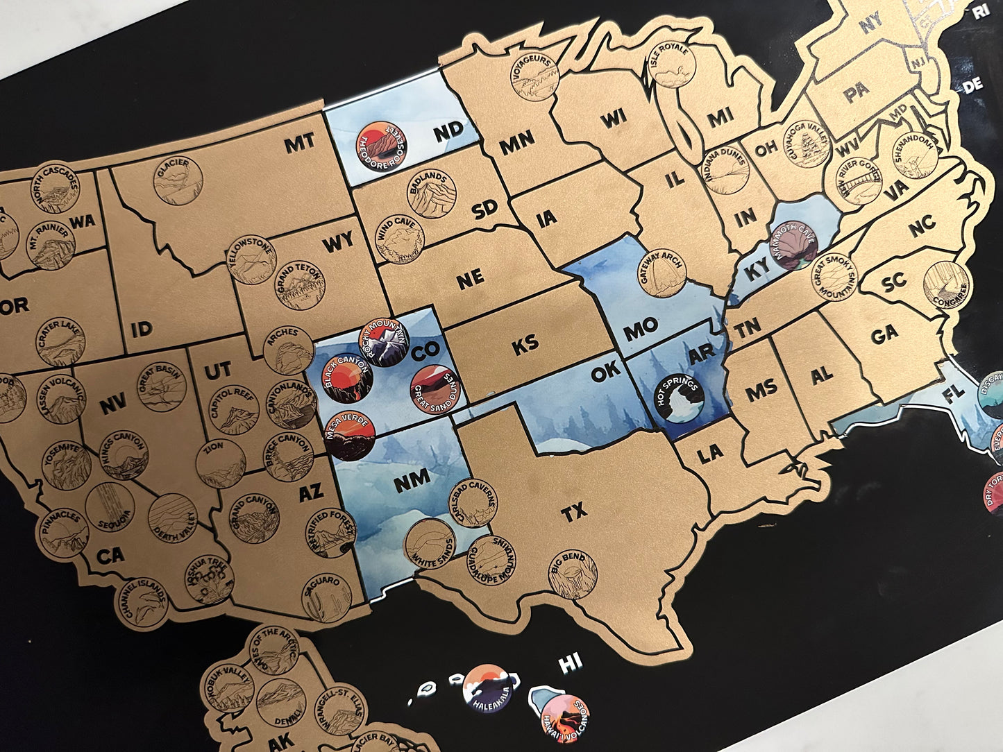 63 National Park and USA scratch off map, Has a black background with a blue watercolor design underneath a gold layer, which scratches off to reveal artwork.