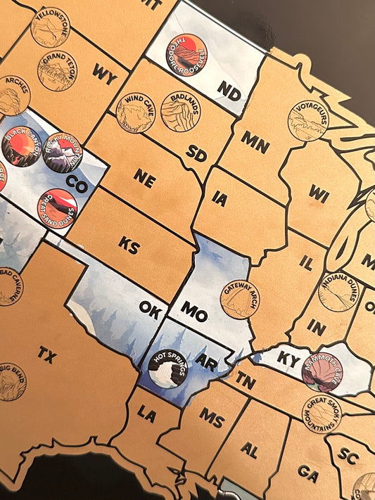 63 National Park and USA scratch off map, Has a black background with a blue watercolor design underneath a gold layer, which scratches off to reveal artwork.