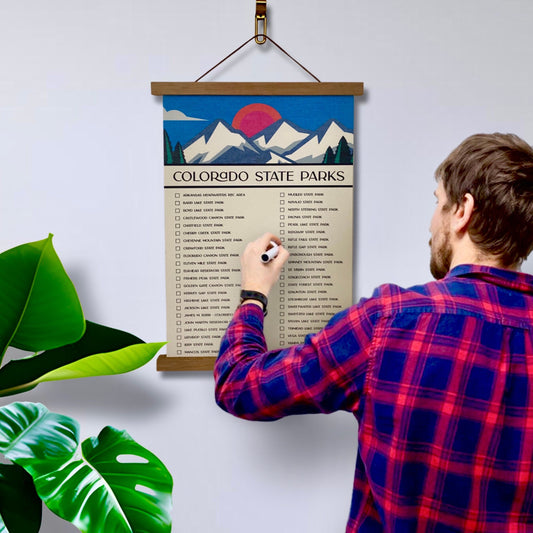 Colorado State Parks Checklist Tracker with marker. Printed on canvas. Has a design on the top with a mountain and sun. Hanging on a wooden frame.