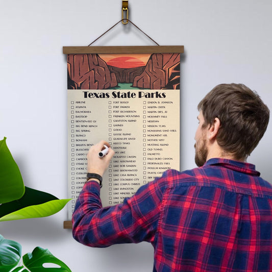 Texas State Parks Checklist with marker. Printed on canvas. Has a design on the top with a sunset and canyon. Hanging on a wooden frame.